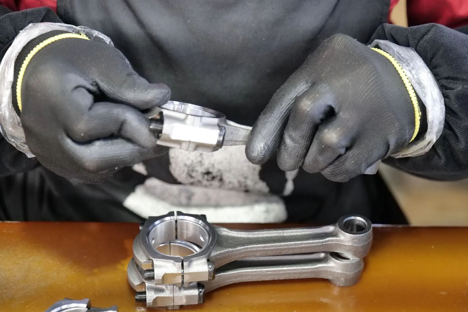 Gloved hands working on a connecting rod above a workbench with two more connecting rods and their bearing shells