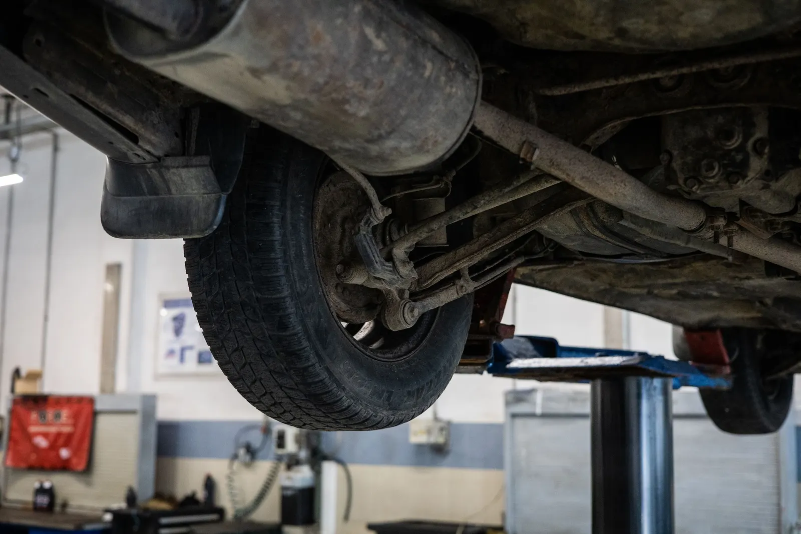 Underside of a vehicle raised on a two-post lift in a repair shop, showing a rear wheel, suspension arms and the exhaust