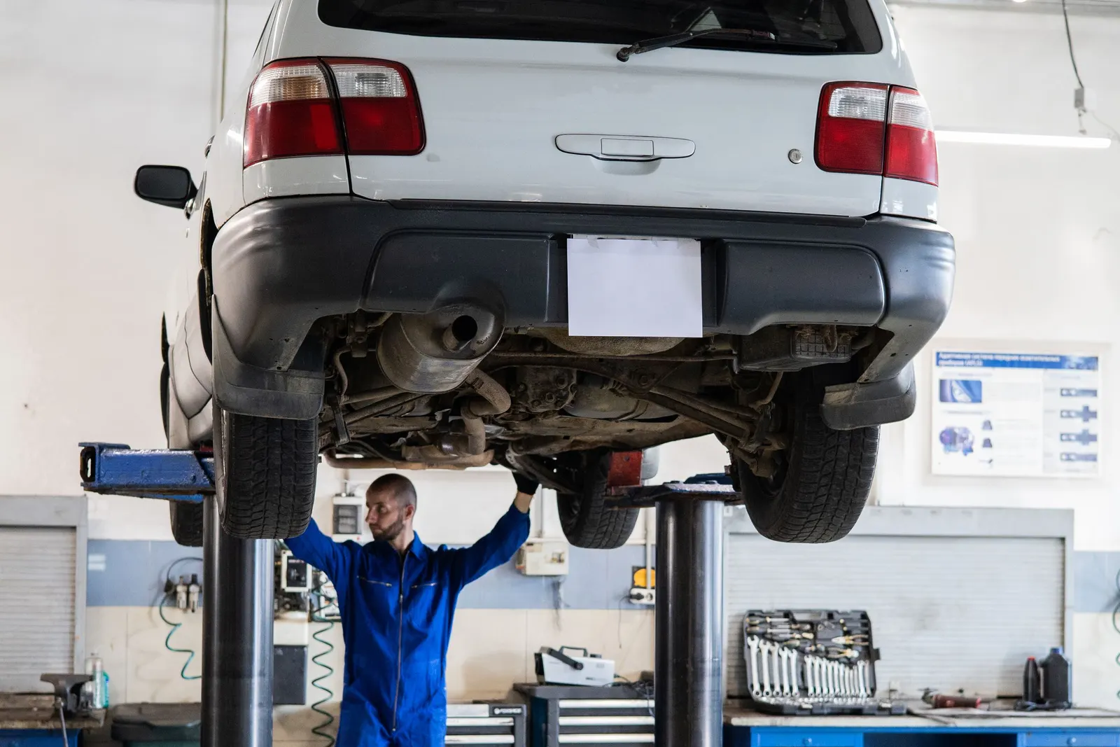 Mechanic standing under a white SUV raised on a two-post lift in a repair shop, inspecting the underside