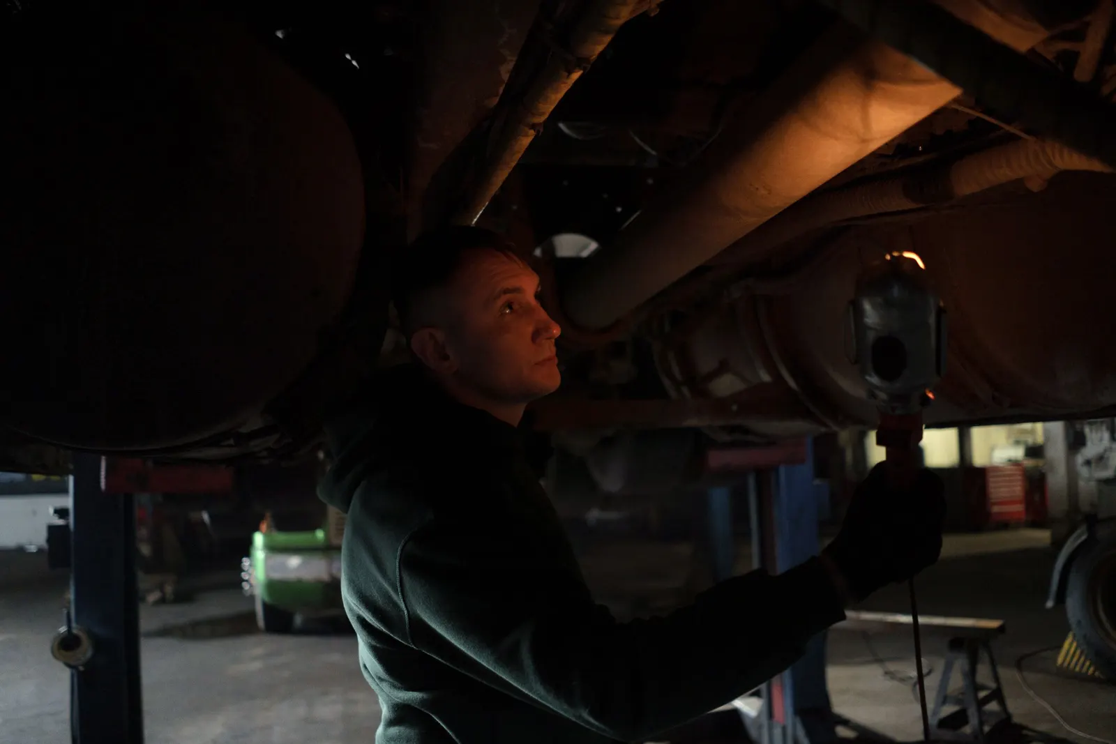Mechanic standing under a raised vehicle holding a work light up to the underside to look for the source of a leak