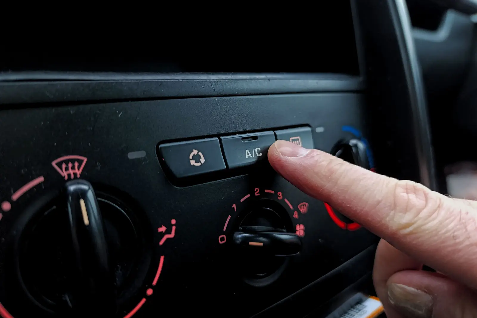 Finger pressing a button next to the A/C button on a car's climate control panel, beside the windshield defrost symbol