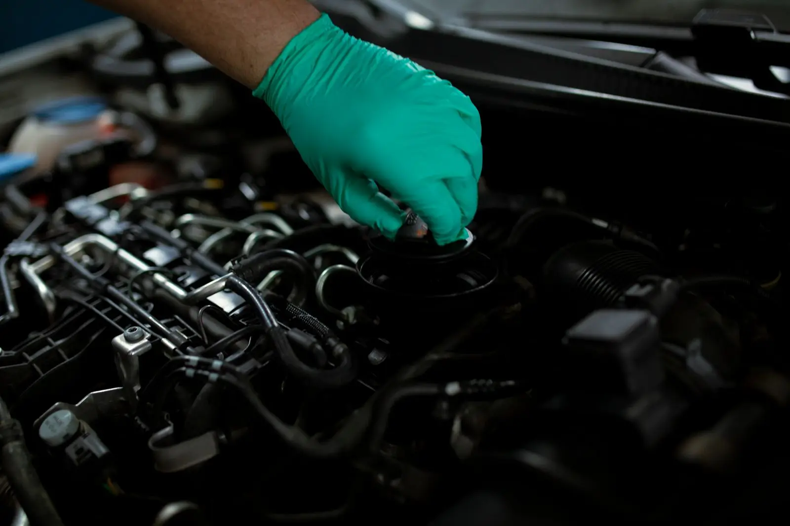 Gloved hand unscrewing the oil fill cap on top of a car engine, with fuel lines and wiring around it
